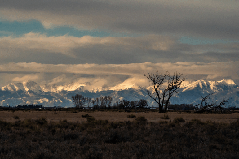 The Sangre de Cristo mountains loom over Colorado’s San Luis Valley. A beautiful landscape of snow covered mountains and white clouds is pictured.