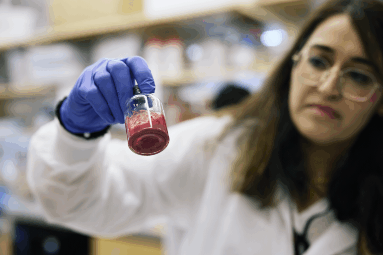 This photo shows a woman holding a vial of red artificial blood. She's wearing a white lab coat and a blue glove.