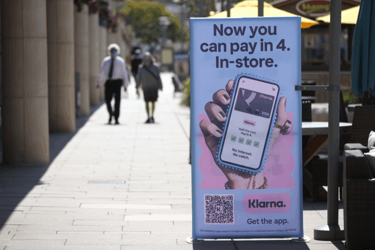 Pedestrians walk toward a sidewalk sign that reads "Now you can pay in 4. In-store" with a picture of a hand holding a cellphone.