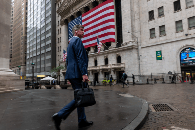 A businessman wearing a dark blue suit walks by the New York Stock Exchange, whose facade is draped with a huge American flag.