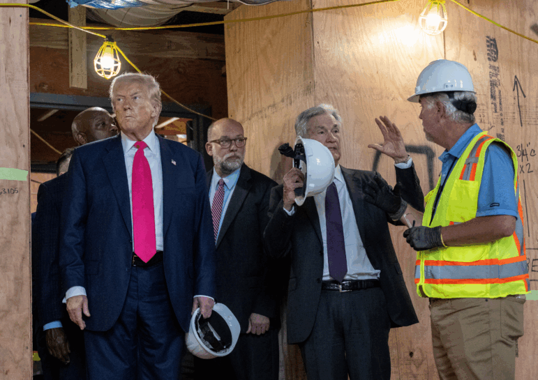 This photo shows President Trump, Federal Reserve Chair Jerome Powell and the director of the Office of Management and Budget, Russell Vought, during a tour of renovations at the Federal Reserve in Washington, D.C. Powell is speaking with the construction manager, who is wearing a fluorescent vest. Wooden boards cover many areas of the building.