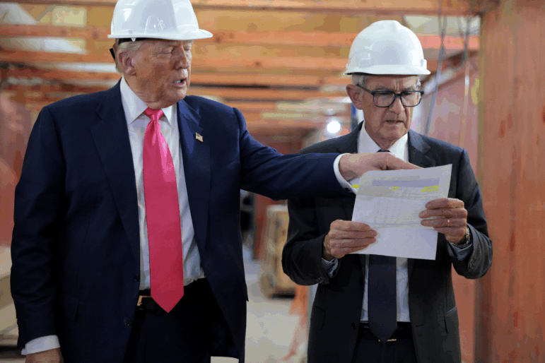 In this photo, Federal Reserve Chair Jerome Powell and President Trump, both wearing suits, ties and hard hats, walk through the central bank's headquarters, which is under renovation, with wooden beams and boards visible. Powell is holding a sheet of paper, and Trump is pointing to something on it.