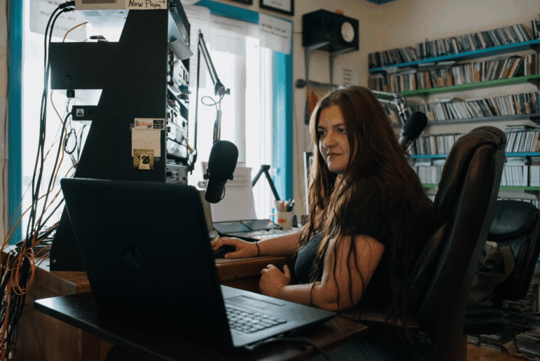 Abby DuFour, WCHG News Reporter and afternoon host of DuFour Du Jour, cues up the next song in her broadcast at station WCHG 107.1 FM, in Hot Springs, Virginia, on Friday, July 18th, 2025. Credit: Kristian Thacker for NPR
