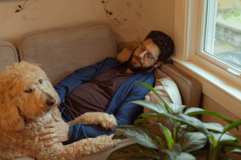 Benjamin Stein-Lobovits, a young man who has dark curly hair, a beard and glasses, lays on the couch petting his dog, Cow, a large, beige poodle mix.
