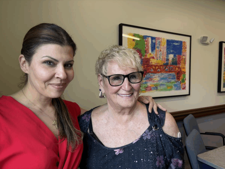 Rachel Nassif has her arm around Roberta Rabinovitz shoulder. The two women are standing in front of a colorful painting and smiling.