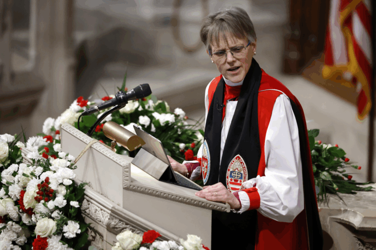 Bishop Mariann Edgar Budde, wearing clerical attire in red, white and black speaks at a podium adorned with flowers.
