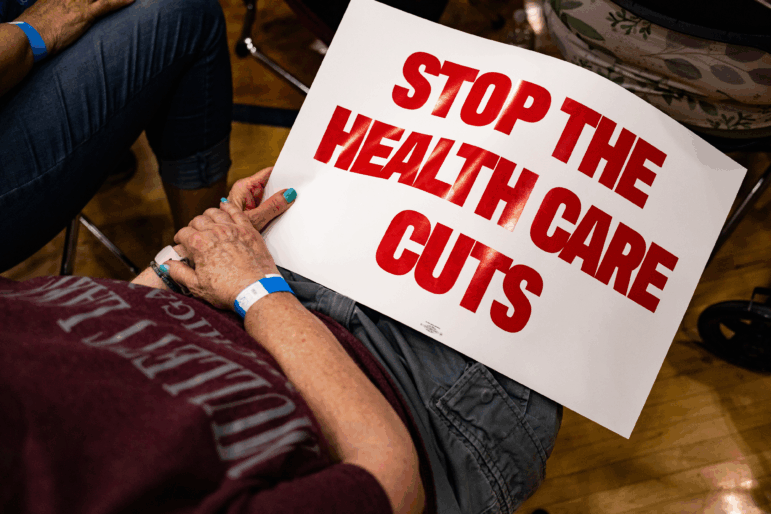An attendee holds a "Stop The Health Care Cuts" sign during a June 6 town hall hosted by Michigan Democrats in Lansing. In July, Congressional Democrats failed to stop Republicans from passing a tax-and-spending bill that mandated that 40 states and the District of Columbia institute work requirements for Medicaid eligibility. Michigan launched a system for verifying work requirements in 2020, but shut it down after a court ruling.