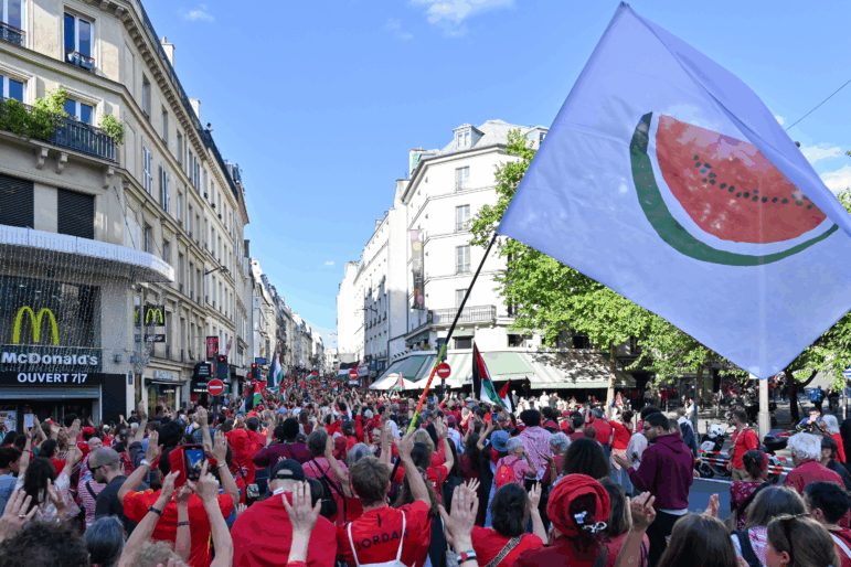 A white flag with a large watermelon slice on it is waved during the Red Line for Gaza protest in Paris on July 8. Most of the protesters are wearing red shirts, and they're walking down a street lined by multistory buildings.