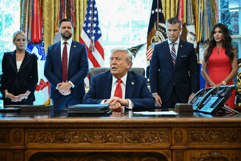 President Trump sits at a wooden desk at the White House as he speaks to the press. Behind him stand (from the left to right side of the frame) Attorney General Pam Bondi, Vice President Vance, Defense Secretary Pete Hegseth and Secretary of Homeland Security Kristi Noem.
