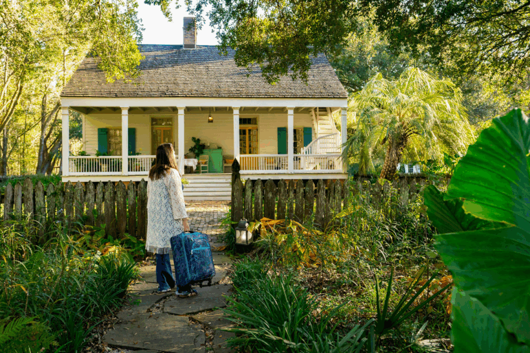 A rearview photograph of a person holding a rolling suitcase as they stand on a stone pathway facing a yellow cottage with a quaint front porch. The person walks along the stone path, which is framed by lush foliage, approaching the house where they will stay as a houseguest.