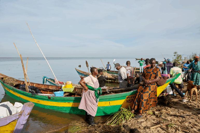 Alice Akinyi (left, hand on hip) and Justine Adhiambo Obura, members of the No Sex For Fish group, stand next to Alice's fishing boat at Nduru Beach. It's one of the few boats that have weathered the storms and floods that have beset the area since 2020.