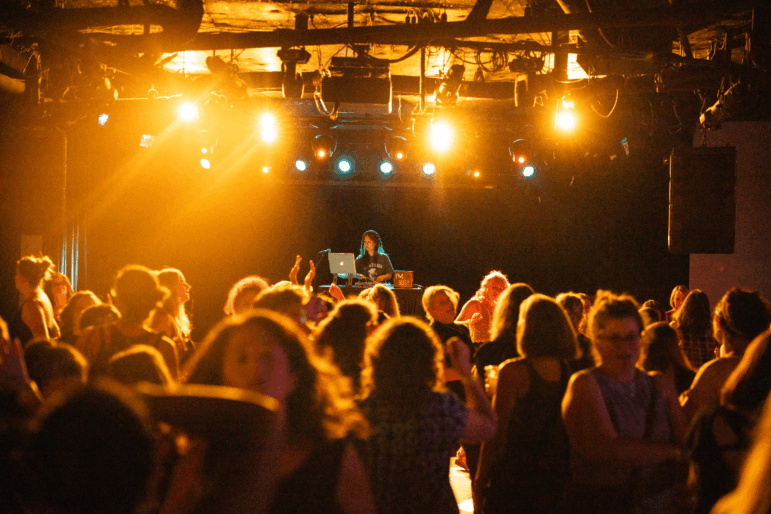 A DJ plays music on stage from a laptop as a crowd of women dance under yellow stage lights.