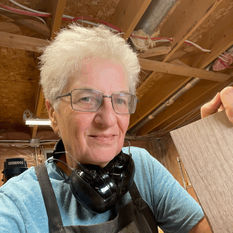 This photo shows Doris Kochanek in her basement workshop in a suburb of Ottawa, Canada. One of her hands is atop a sheet of wood that's standing on one of its edges.