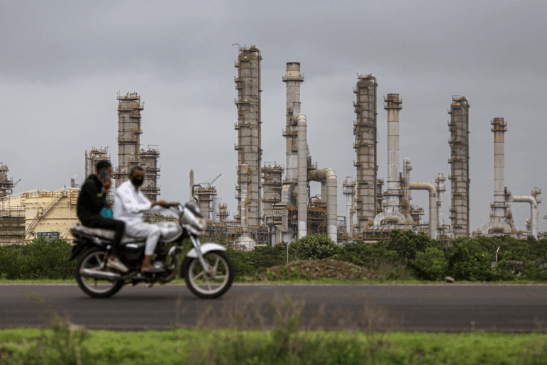 Reliance Industries' oil refinery in Jamnagar, India, is a network of metal towers, pipes and tanks in this photo. In the foreground, two men ride a motorbike on a road.