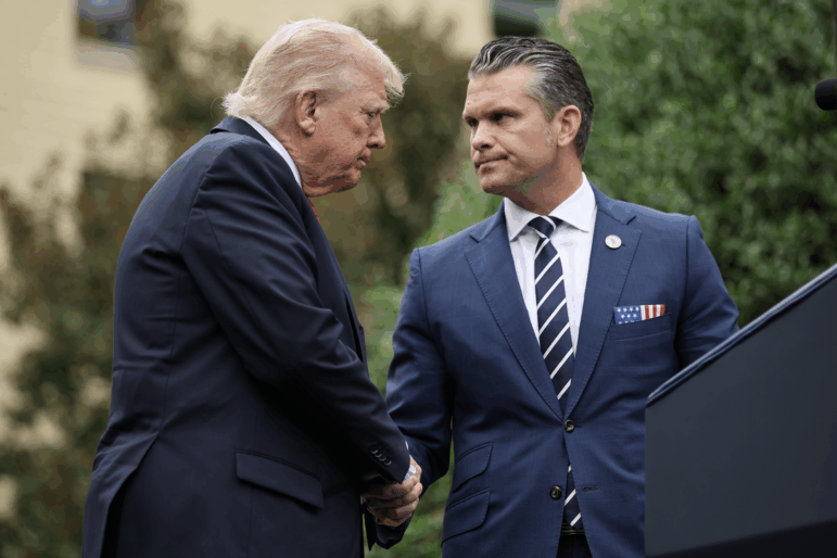 President Trump and Defense Secretary Pete Hegseth shake hands. They are standing outdoors and are wearing blue suits. Trump is on the left side of the frame, and Hegseth is on the right side of the frame.