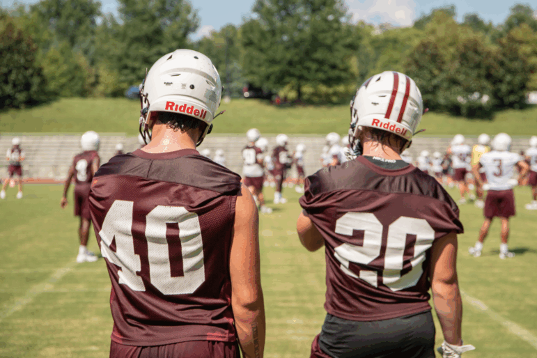 Two players with their backs to the camera watch the players on the football field from the sidelines. One is wearing jersey No. 40 and one wears No. 20. Both are wearing their football helmets.