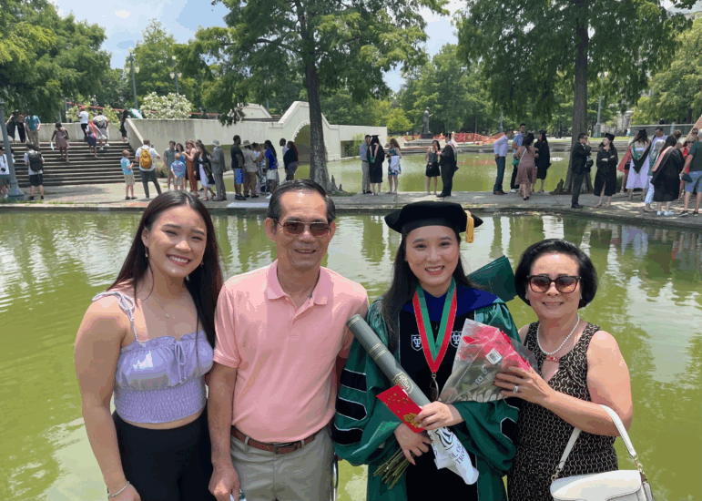 NhuNgoc Pham is shown in a cap and gown, signifying her doctorate. She holds flowers and is standing in front of a fountain with her family.