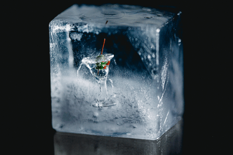 A close-up photograph of a miniature martini glass frozen inside of an ice cube.