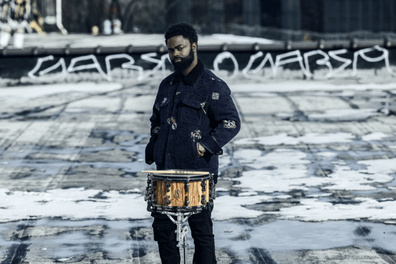 drummer marcus gilmore stands on a rooftop. he wears a dark blue denim jacket. his hands are in his pocket. he stands in front of a snare drum with drum sticks laying across it.