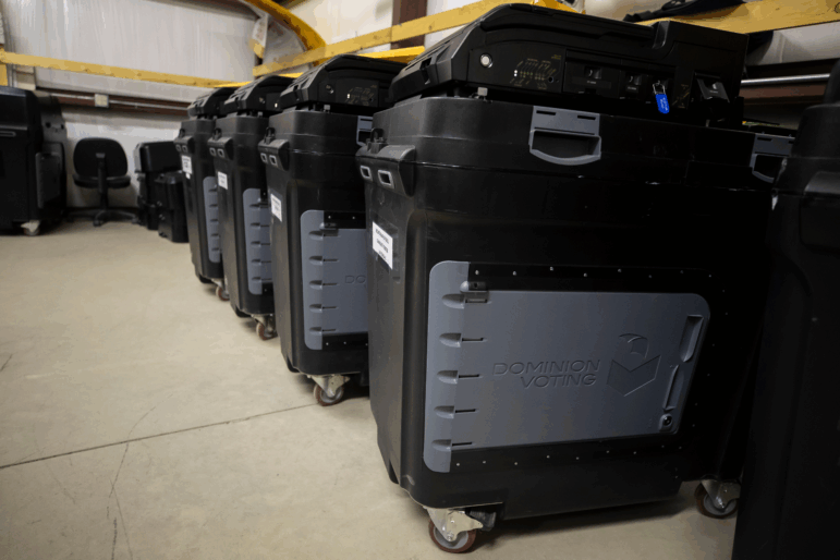 Dominion Voting Systems ballot-counting machines, which have a black plastic exterior and wheels on the bottom, are lined up inside a Torrance County warehouse in Estancia, N.M., on September 29, 2022.