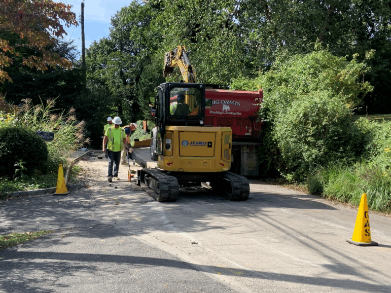A gas pipeline construction crew in Wyncote, Pennsylvania, replaces older pipes that are prone to leaking climate-heating methane. Men in hard hats work alongside construction equipment on a street that's lined with foliage.