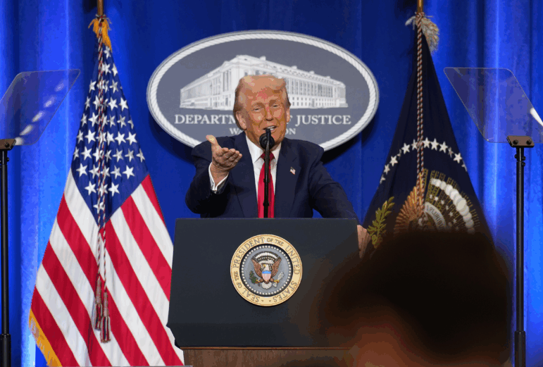 Standing behind a lectern equipped with a microphone, President Trump gestures with his right hand outstretched while speaking at the Justice Department on March 14, 2025.