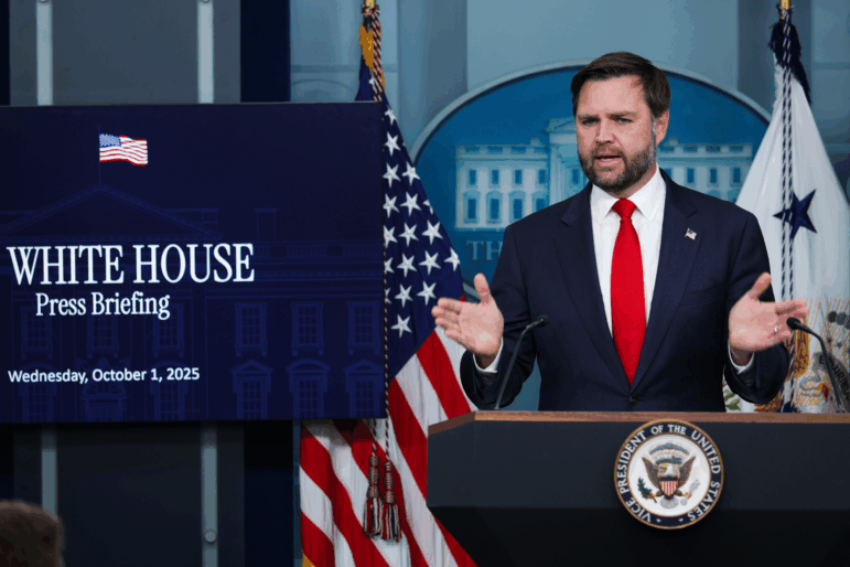 Vice President JD Vance speaks at a podium next to a sign that says "White House Press Briefing, Wednesday, October 1, 2025." An American flag is behind him and the presidential seal is on the podium.