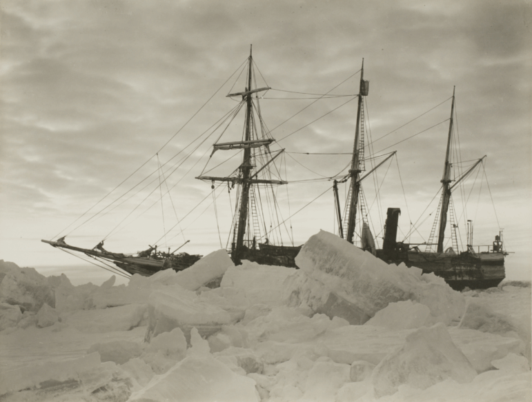 This black-and-white photo shows the ship Endurance in the winter of 1915. Large pieces of sea ice are in the foreground, with the three-masted ship behind the ice.