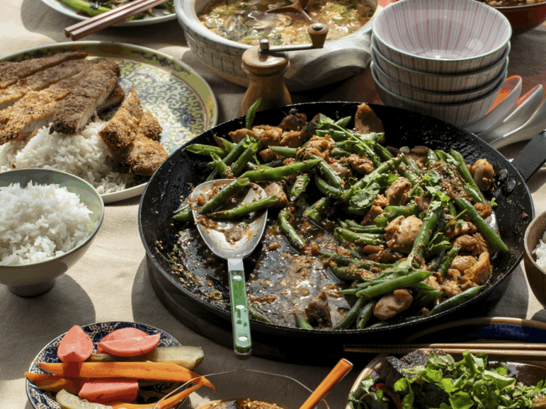 This photo shows a green bean and chicken stir-fry, with sauce and seasonings, in a black pan. It is surrounded by other dishes that include vegetables and rice.