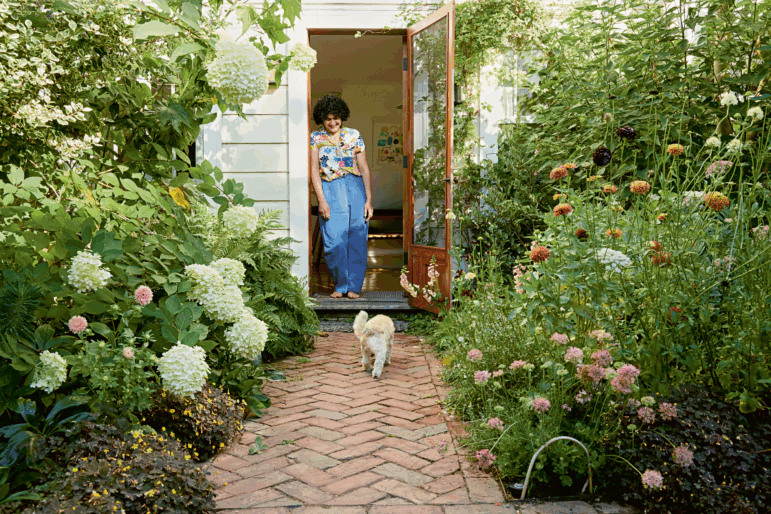 A photograph shows chef and cookbook author Samin Nosrat standing in the doorway of a house, colorful art hangs on the wall behind her, the glass door opens to her right, and she's surrounded by tall, leafy hydrangeas, allium, ferns, and other plants on either side. Samin is barefoot, wearing a colorful button-up shirt, leaning casually against the doorframe, and smiling down at a small white dog walking toward her on a brick pathway.