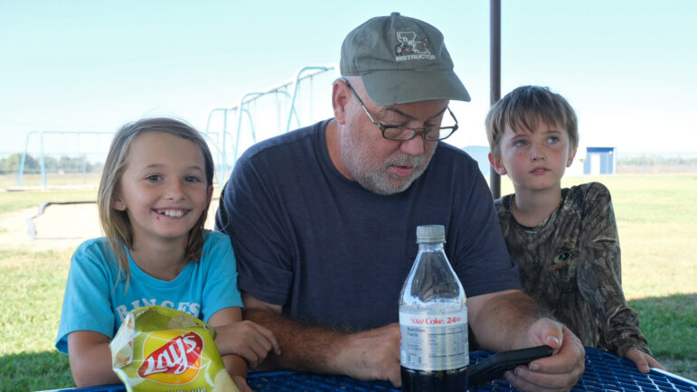 Penelope Hull, her grandfather Randy Ogles, and brother Lucien Hull sit at a picnic bench outside Holly Ridge Elementary School in Holly Ridge, Louisiana, on Friday, October 17, 2025.