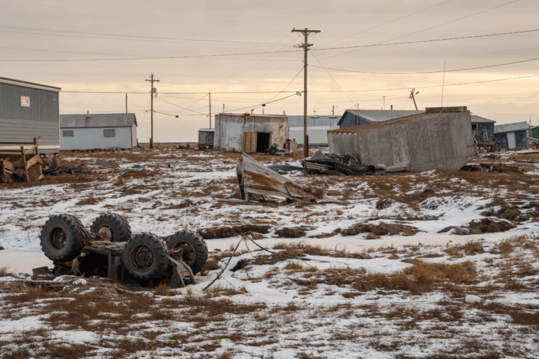 A shed-like building and an all-terrain vehicle lie overturned upside down in Kwigillingok, Alaska, in late October. The ground is partially covered by snow. In the background stand a few small, one-story buildings. Power lines supported by poles crisscross the area.