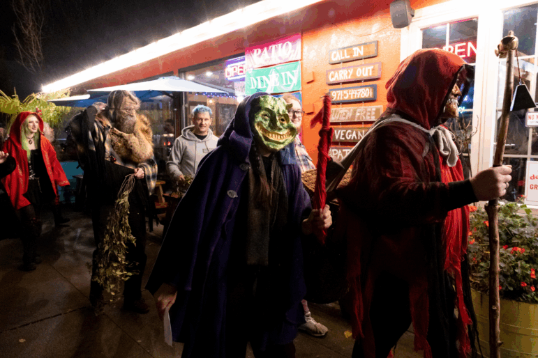 People dressed in creepy costumes and masks walk in a procession. The person in front is wearing a red hood and carrying a staff with a bell, and the next person in line is wearing a green monstrous mask with a wide, toothy smile.