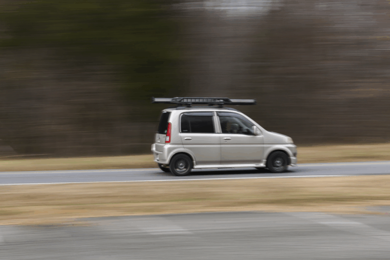 A small, gray, four-door Honda Life kei car zips by on a road.