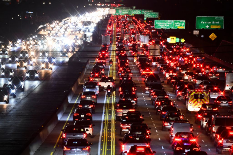 Multiple lanes of cars and other vehicles crawl slowly in rush-hour traffic on Interstate 405 in Los Angeles in March 2022. The scene is after dark, and the vehicles have their headlights and taillights turned on.