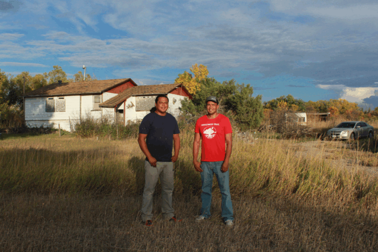 Lonny and Teyon Fritzler stand outside their childhood home. Plywood covers the windows of the white house, and tall gold-colored grass is high in the yard between the brothers and the house.
