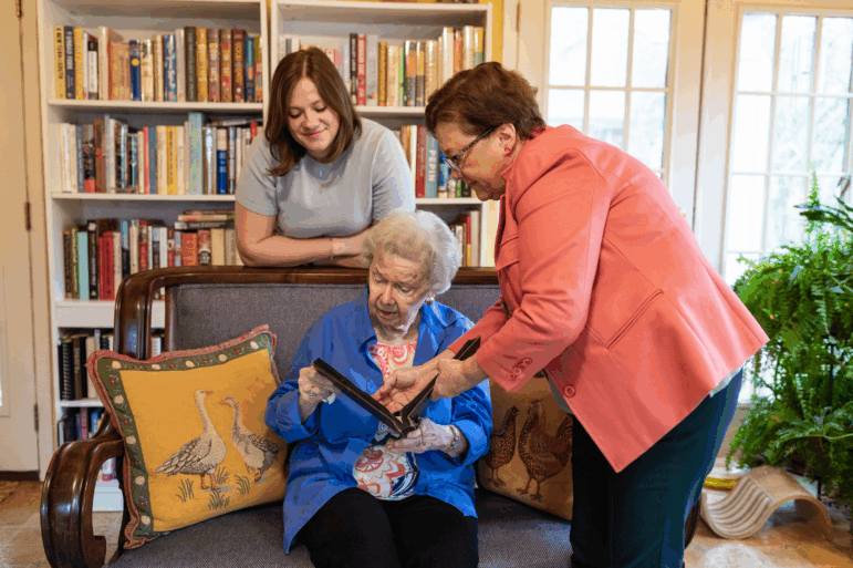 Caroline Brown (left) and Cynthia Briscoe Brown look at family photo album with their grandmother and mother Barbara Briscoe (seated) in Atlanta. Caroline, 33, is wearing a gray t-shirt and standing behind a vintage couch. Cynthia, 65, is wearing a pink blazer and holding a photo album while Barbara, 93, is seated. Barbara is wearing a blue shirt over a pink patterned shirt.