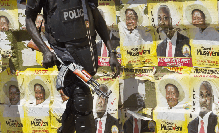 A Ugandan riot policeman carrying a large firearm with a shoulder strap walks past a wall plastered with campaign posters for longtime President Yoweri Museveni.