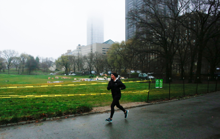 A jogger is seen in warm winter running clothes on a paved path in front of a wide open, green lawn. Buildings are in the background.