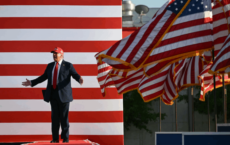Wearing a suit and red baseball cap, President Trump stands onstage with arms outstretched to deliver remarks at the Iowa State Fairgrounds on July 3, 2025. Multiple American flags wave on flagpoles on the right side of the frame.