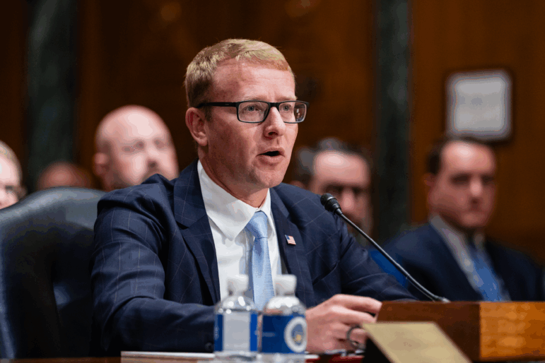 A man in a dark suit, white shirt and blue tie, with short hair and wearing glasses,  sits at a table in a congressional hearing room and speaks into a microphone.