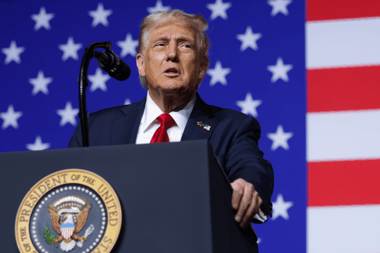 President Trump, wearing a suit with red tie, stands behind a lectern that's equipped with a microphone and that has the seal of the U.S. president on the front of it. The backdrop behind him is the U.S. flag.