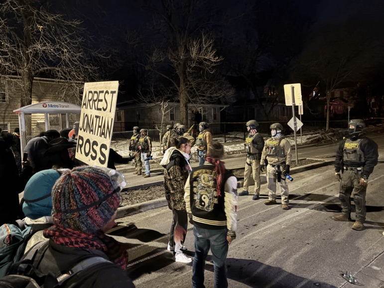 Demonstrators stand facing front federal immigration agents who are masked and wearing helmets. One demonstrator holds a large sign that reads, "Arrest Jonathan Ross."