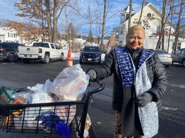 Marilyn Vargas, wearing a coat and scarf, stands in a parking lot, with one hand holding onto a shopping card filled with groceries, most in white plastic bags. Behind her are parked cars and snow drifts.