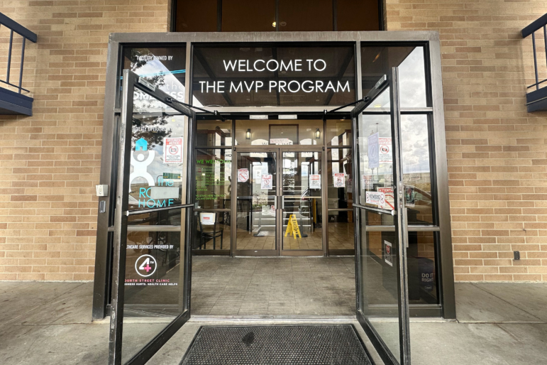 This photo shows the double glass doors of the Medically Vulnerable People shelter in Sandy, Utah. The building's exterior walls are brown bricks. Above the glass doors are the words "WELCOME TO THE MVP PROGRAM."