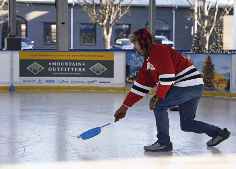 A man wearing a red hockey jersey and a red plaid winter hat tosses a blue cast iron skillet on an ice rink.