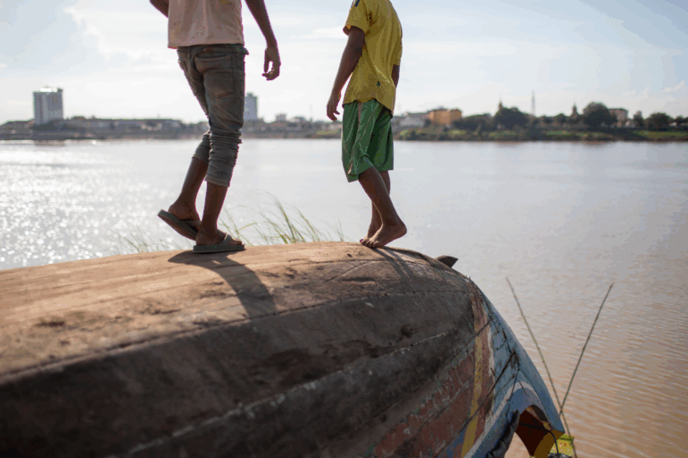 Two children stand on top of an overturned boat along a bank of the Mekong Delta.