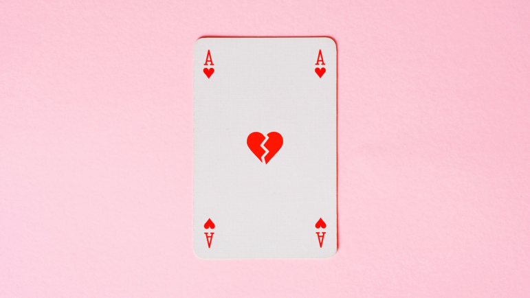 A photograph of an ace-of-hearts playing card shows a broken heart in the center of the card, symbolizing being unlucky in love and having relationship issues. The card is photographed straight on, on a light pink background.