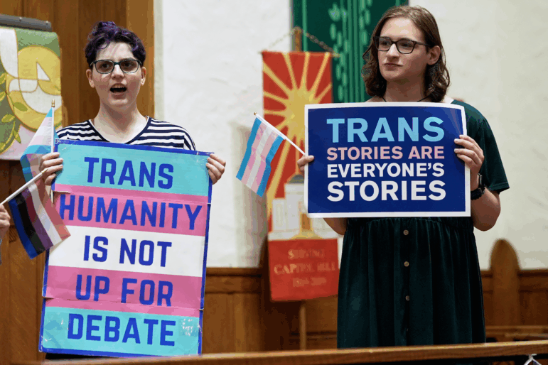 Two people hold signs as they stand near the front of a church. One reads: "Trans humanity is not up for debate." The other says: "Trans stories are everyone's stories."