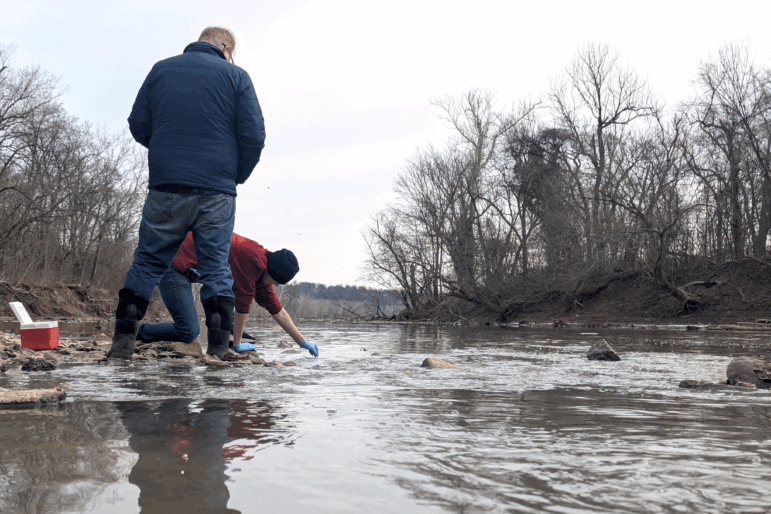 Two men take water samples from the Potomac River in Maryland in January. One man is wearing blue disposable gloves and is crouching down to reach into the river to collect a sample.